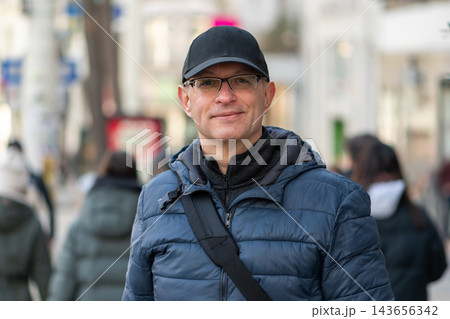 Smiling man in cap and glasses standing on busy street Smiling man in cap and glasses standing on busy street 143656342