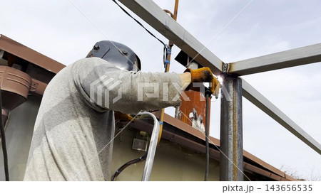 Manual laborer in protective mask weld iron detail on carport frame. Professional young repairman with welding machine working at construction. Male welder operating with metal at the site. Close up Manual laborer in protective mask weld iron detail on carport frame. Professional young repairman with welding machine working at construction. Male welder operating with metal at the site. Close up 143656535