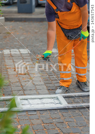 Road worker paints pedestrian crossing lines on cobblestone street using spray equipment during road maintenance. Road worker in orange uniform applies white marking paint for pedestrian safety 143657543
