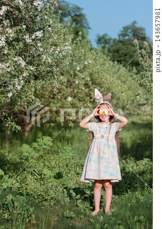 Young girl wearing bunny ears holding colorful Easter eggs over eyes in blooming orchard under blue sky Young girl wearing bunny ears holding colorful Easter eggs over eyes in blooming orchard under blue sky 143657981