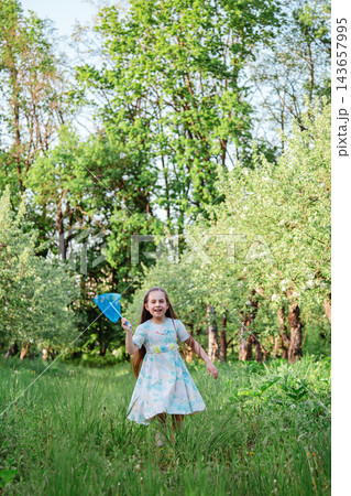 Happy little girl in floral dress playing with blue butterfly net outdoors in vibrant green orchard Happy little girl in floral dress playing with blue butterfly net outdoors in vibrant green orchard 143657995