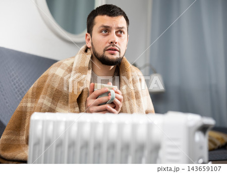 Man with a blanket on his shoulders with cup near electric heater Man with a blanket on his shoulders with cup near electric heater 143659107