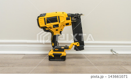 Yellow cordless fastening tool standing upright on a wood floor against a plain wall in an indoor home setting Yellow cordless fastening tool standing upright on a wood floor against a plain wall in an indoor home setting 143661071