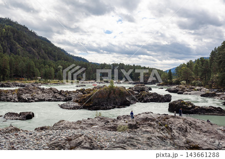 Scenic view of a river with rocky banks, surrounded by dense green forests and mountains under a cloudy sky, featuring people along the shore Scenic view of a river with rocky banks, surrounded by dense green forests and mountains under a cloudy sky, featuring people along the shore 143661288