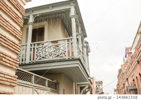 Ornate gray balcony with patterned railing and hanging lights projects between historic brick facades, overlooking a narrow tbilisi street Ornate gray balcony with patterned railing and hanging lights projects between historic brick facades, overlooking a narrow tbilisi street 143661338
