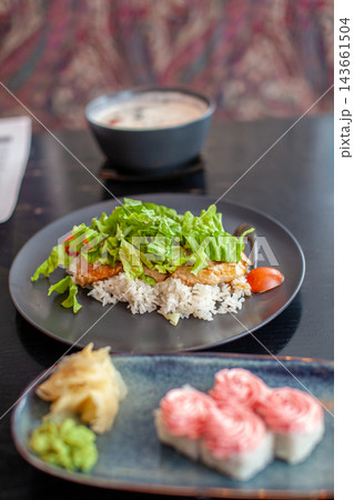 Plate features rice topped with grilled chicken, fresh greens, and cherry tomatoes, alongside sushi and miso soup in a restaurant environment 143661504