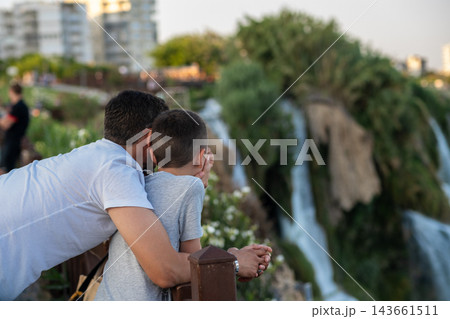 A father and his son closely observe the beautiful waterfall amidst greenery, capturing a shared moment of awe in the park during late afternoon. A father and his son closely observe the beautiful waterfall amidst greenery, capturing a shared moment of awe in the park during late afternoon. 143661511