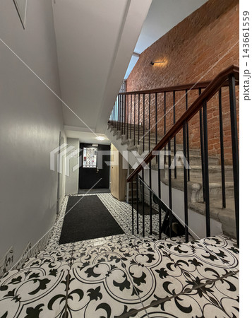 Interior view of a staircase featuring decorative tile flooring and a wooden railing, with an exposed brick wall and a black door at the top Interior view of a staircase featuring decorative tile flooring and a wooden railing, with an exposed brick wall and a black door at the top 143661559