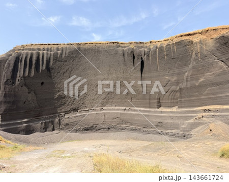 Broad volcanic tuff cliff displays dark vertical erosion channels, horizontal strata, sandy ground, sparse grasses, and a bright blue sky above 143661724