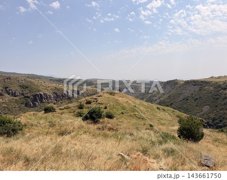 Dry golden grass covers rolling hills above a rugged rocky valley, with scattered shrubs, distant ridges, and a bright blue sky overhead Dry golden grass covers rolling hills above a rugged rocky valley, with scattered shrubs, distant ridges, and a bright blue sky overhead 143661750