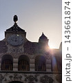 Detailed stone facade features multiple arched wooden doors, sculpted floral reliefs, pointed towers, and bright sunlight behind the historic structure 143661874