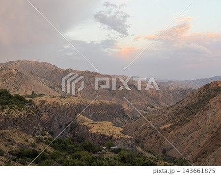 Rugged mountain valley displays layered dry slopes and distant ridges, with scattered green trees and pastel clouds beneath softly lit sky 143661875