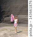 Woman in a white top and pink shorts holds matching patterned fabric before a layered dark rock wall and open gravel ground 143661876