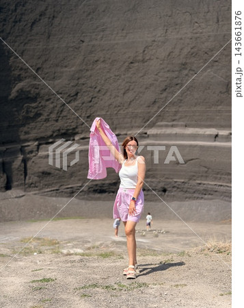 Woman in a white top and pink shorts holds matching patterned fabric before a layered dark rock wall and open gravel ground Woman in a white top and pink shorts holds matching patterned fabric before a layered dark rock wall and open gravel ground 143661876