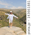 Woman in white clothing and cap stands with arms extended on a concrete overlook, facing a broad dry mountain valley beneath scattered clouds 143661881