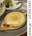Oval baked bread holds melted cheese, sunny egg yolk, and butter on a ceramic plate, with salad remnants and cutlery nearby 143661885