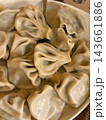 Uncooked folded dumplings with twisted pleats and smooth glossy dough fill a shallow bowl, viewed closely beside a wooden surface 143661886