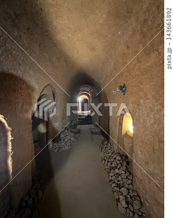 Dim underground passage with rough earthen walls, stacked stones, arched openings, and warm light visible through doorways in a historic subterranean interior 143661892