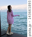 Woman wearing pink striped clothing stands on a dark wooden deck beside blue ocean water, with distant clouds and gentle waves beyond 143661893