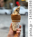 Hand holds a chocolate and vanilla soft serve cone topped with nuts and wafer pieces, served in a patterned paper cup outdoors 143661898