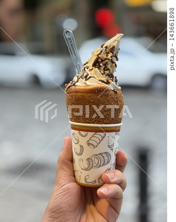 Hand holds a chocolate and vanilla soft serve cone topped with nuts and wafer pieces, served in a patterned paper cup outdoors 143661898