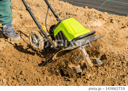 In a garden setting, an electric tiller is working the soil to prepare it for planting. The action of an electric tiller breaking up dirt in a garden plot under bright sunlight. In a garden setting, an electric tiller is working the soil to prepare it for planting. The action of an electric tiller breaking up dirt in a garden plot under bright sunlight. 143662719