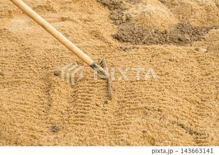 A rake is seen leveling fine sand at a construction site, ensuring an even surface. The focus is on the meticulous action of smoothing the sand for landscaping purposes. 143663141