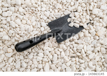 A compact black hand trowel rests on decorative white gravel stones, creating a visually appealing garden scene that highlights landscaping tools and maintenance equipment. A compact black hand trowel rests on decorative white gravel stones, creating a visually appealing garden scene that highlights landscaping tools and maintenance equipment. 143663145