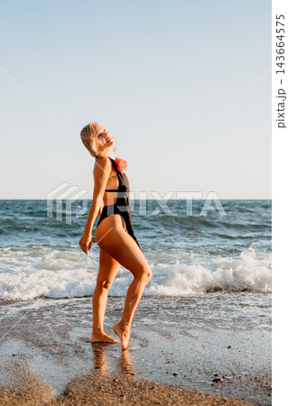 Blonde woman in black swimsuit standing by ocean waves during sunset golden hour Blonde woman in black swimsuit standing by ocean waves during sunset golden hour 143664575