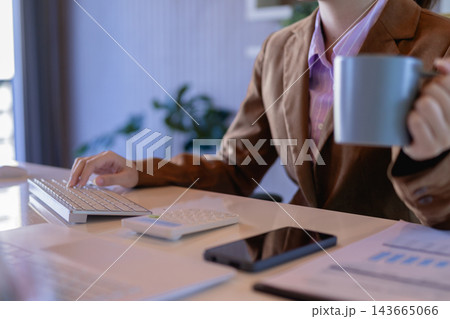 Businesswoman typing keyboard while holding coffee mug at office desk with calculator and phone Businesswoman typing keyboard while holding coffee mug at office desk with calculator and phone 143665066