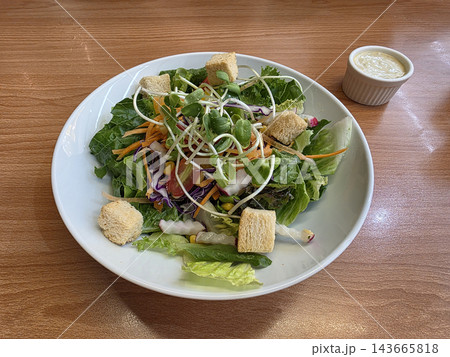 A fresh garden salad with mixed greens, croutons, and sunflower sprouts in a white bowl, served with creamy dressing on a wooden table. A fresh garden salad with mixed greens, croutons, and sunflower sprouts in a white bowl, served with creamy dressing on a wooden table. 143665818