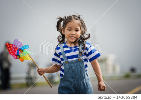 happy preschool child girl walking on road while holding a colorful wind turbine 143665884