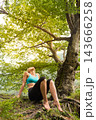 Woman sittLow-angle view of an athletic woman in sports wear leaning back on tree roots and looking up, relaxing under the lush green canopy of a large forest tree 143666258
