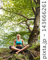 Athletic young woman in sports wear practicing yoga and meditating in lotus pose under the canopy of a large green tree in a serene forest setting. 143666261