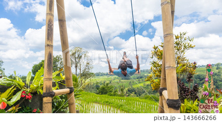 Happy sporty male traveller swinging on wooden swing, enjoying summer vacation in pristine green nature at Jatiluwih rice terraces on Bali, Indonesia. Freedom and life enjoying concept. Happy sporty male traveller swinging on wooden swing, enjoying summer vacation in pristine green nature at Jatiluwih rice terraces on Bali, Indonesia. Freedom and life enjoying concept. 143666266