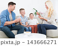 A cheerful lifestyle photo of a family of four sitting on a couch in their living room, playing a card game together. The mother, father, and two young sons are holding playing cards 143666271