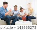 A cheerful lifestyle photo of a family of four sitting on a couch in their living room, playing a card game together. The mother, father, and two young sons are holding playing cards 143666273