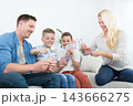 A cheerful lifestyle photo of a family of four sitting on a couch in their living room, playing a card game together. The mother, father, and two young sons are holding playing cards 143666275