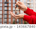 Woman Showing New Home Key With House-Shaped Keychain Outside Residential Apartment Building. First Home Purchase, Homeownership, Mortgage, Real Estate Closing And Moving In Concept. 143667840
