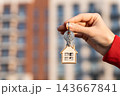 Woman Displaying New Home Key With House-Shaped Keychain Against Residential Apartment Building. Real Estate Purchase, First Home Ownership, Mortgage, Moving In And Housing Advertising Concept. 143667841