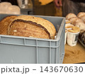 Fresh artisan loaves arranged in gray plastic crate at bakery market with pastries and baker in background Fresh artisan loaves arranged in gray plastic crate at bakery market with pastries and baker in background 143670630