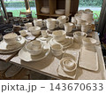 Handcrafted white ceramic tableware arranged on a display table beside sunlit windows and greenery Handcrafted white ceramic tableware arranged on a display table beside sunlit windows and greenery 143670633