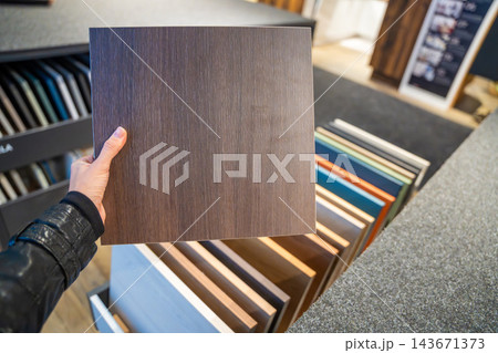 A hand holds a brown wood grain panel above rows of material samples in a showroom. Comparing surface finishes helps plan furniture and interior renovation. 143671373