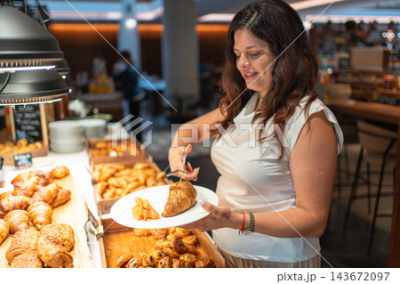 Woman chooses croissant, pancakes, salmon and cheese from a hotel breakfast buffet. Self service breakfast offers guests a variety of food during travel 143672097