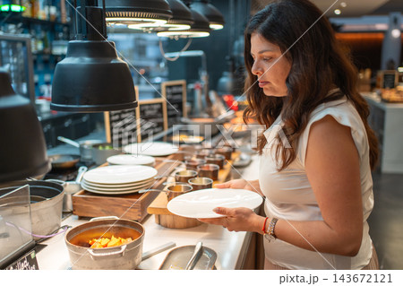 Woman chooses hot food while holding a plate at a hotel breakfast buffet. Morning dining scene represents self service and hospitality for travelers Woman chooses hot food while holding a plate at a hotel breakfast buffet. Morning dining scene represents self service and hospitality for travelers 143672121