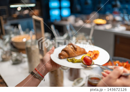 Woman chooses croissant, pancakes, salmon and cheese from a hotel breakfast buffet. Self service breakfast offers guests a variety of food during travel 143672126