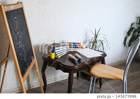 Art space includes a chair, chalkboard, easel, and plant in a room with plain walls and wooden floor Art space includes a chair, chalkboard, easel, and plant in a room with plain walls and wooden floor 143673996