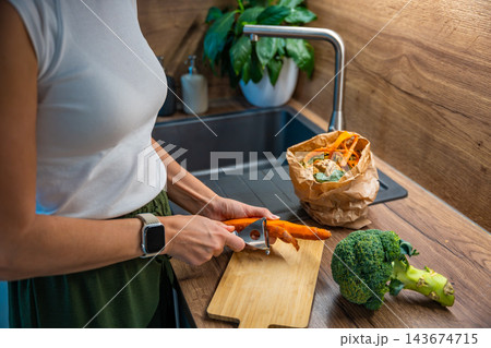A woman peels a carrot beside broccoli and a paper bag of food scraps. Collecting vegetable peelings supports composting during meal preparation. A woman peels a carrot beside broccoli and a paper bag of food scraps. Collecting vegetable peelings supports composting during meal preparation. 143674715