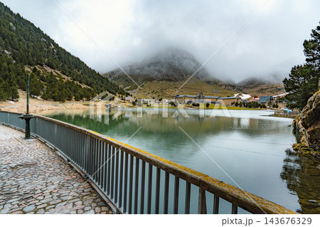 Cobbled footbridge with railing over a green mountain lake with fog covered peak in Vall de Nuria 143676329