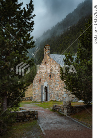 Small stone chapel with bell gable among pine trees below misty mountain slopes in Vall de Nuria 143676336